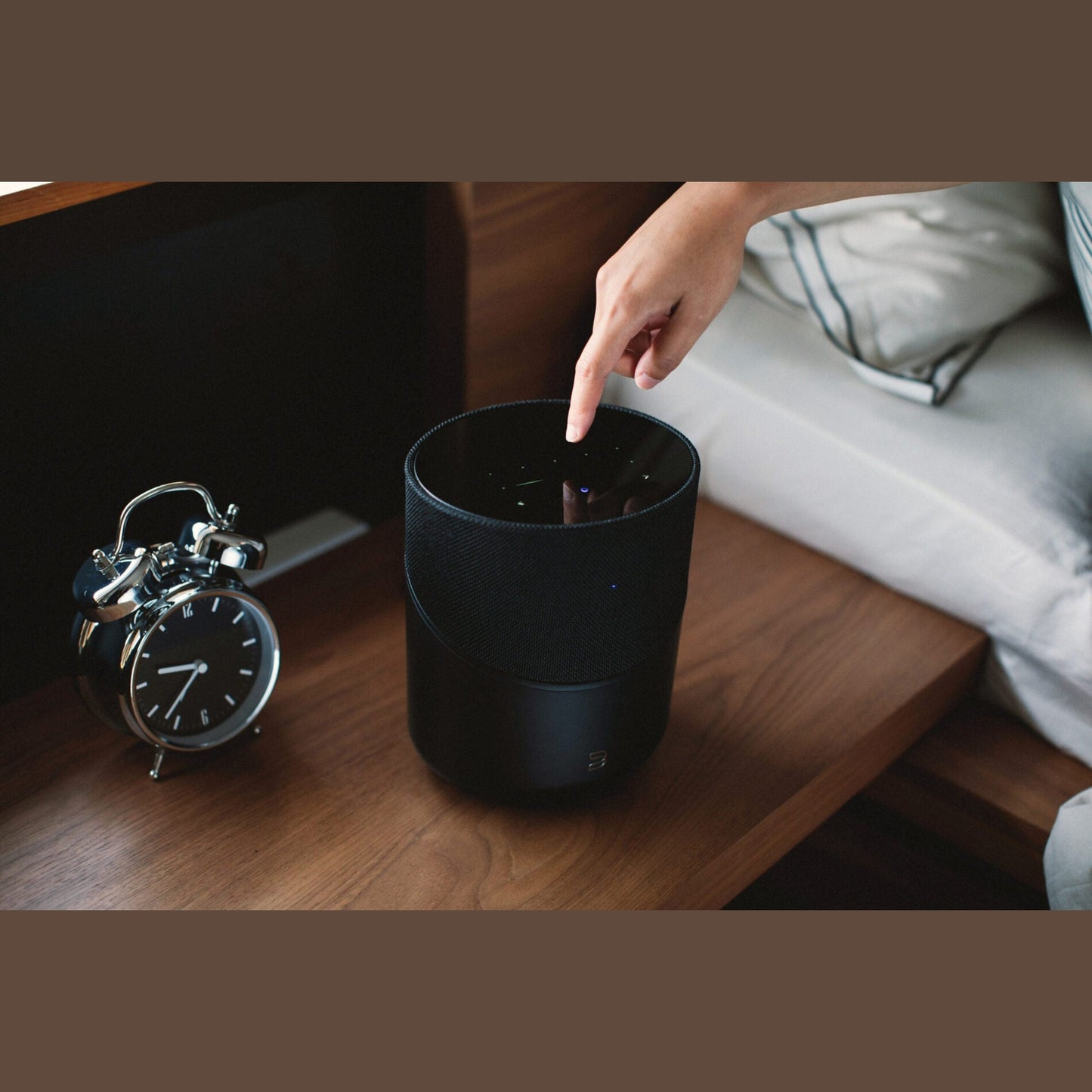 Bluesound Pulse M Streaming-Lautsprecher Schwarz Lifestyle auf dem Nachttisch neben Wecker, Hand am Bedienfeld